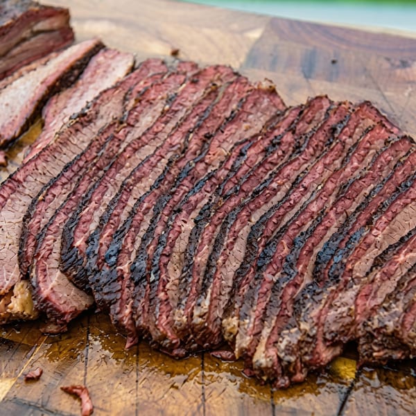 Close-up of thinly sliced, smoked brisket with a dark bark on a wooden cutting board.
