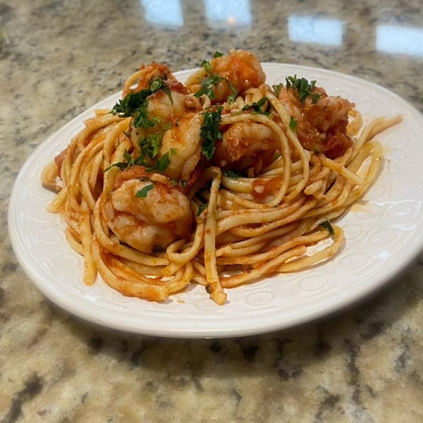 A white plate of shrimp scampi with linguine pasta and parsley on a granite countertop.