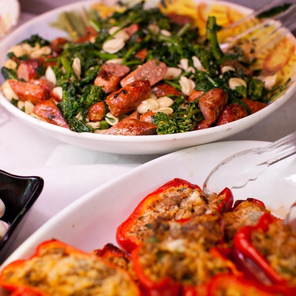 A white bowl of sausage, broccoli rabe, and beans, with stuffed red peppers in the foreground.