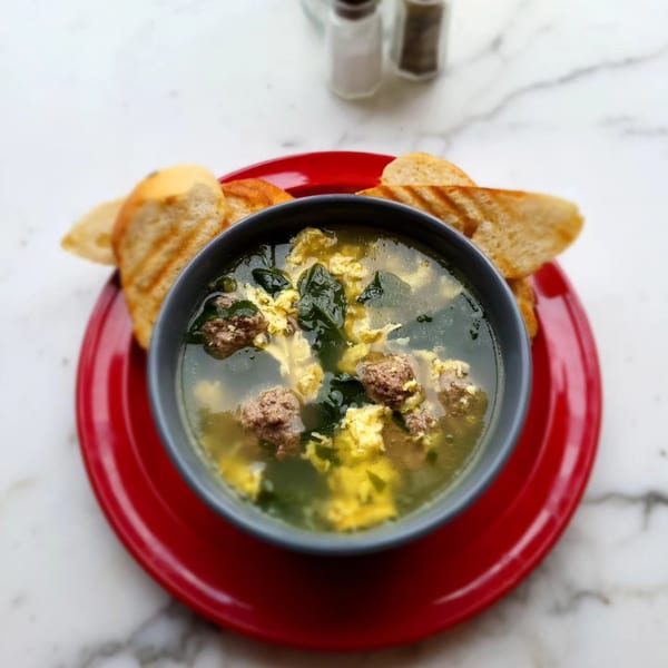 A bowl of clear soup with meatballs, green vegetables, and scrambled egg, served with toasted bread.